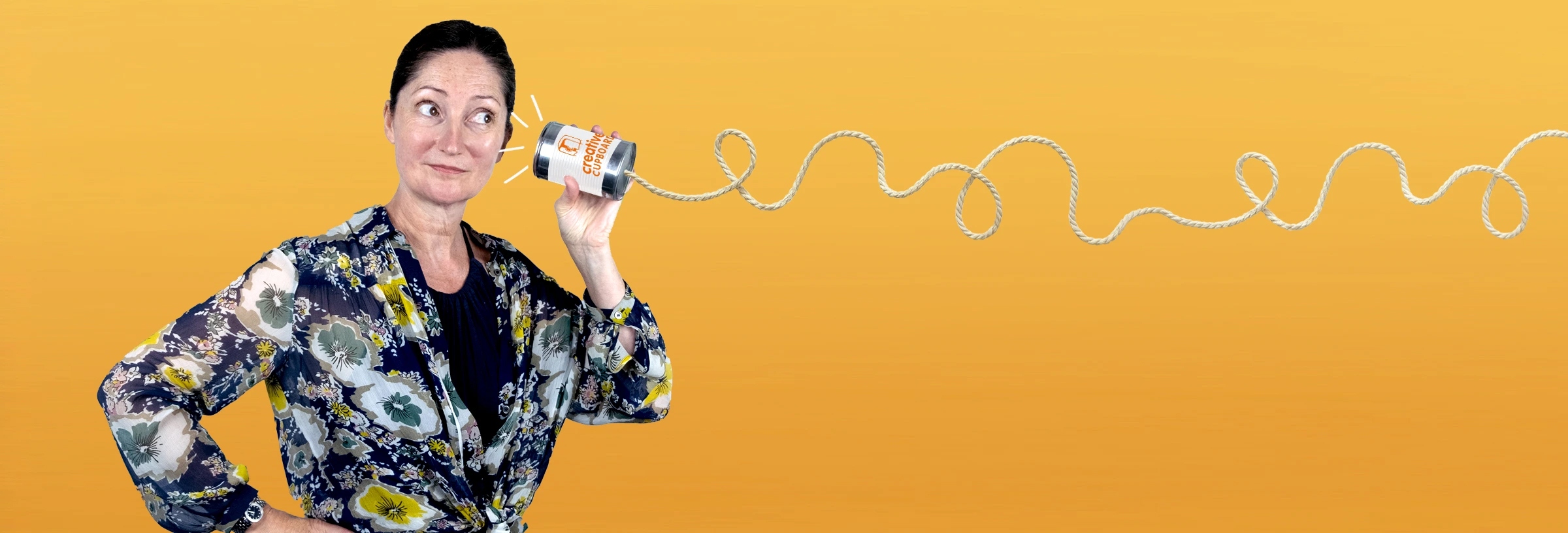 A woman holding a tin can attached to a piece of string to her ear in front of a yellow background
